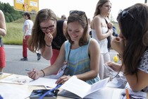 A girl writing something on a sheet. A second girl is looking over her shoulder and stifling a laugh.