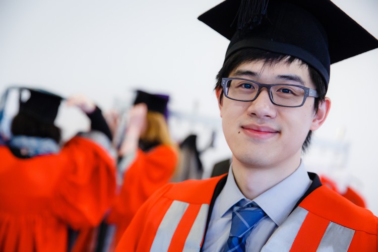 A graduate with glasses wearing a motarboard looking directly into the camera