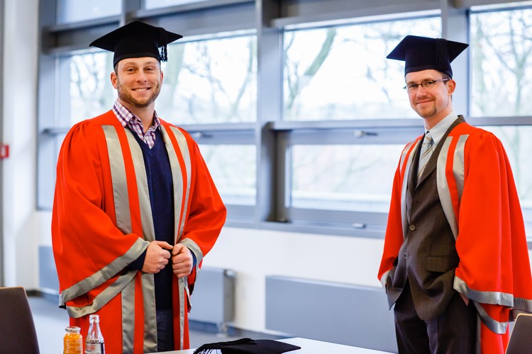 Two graduate in motarboards and gown, standing slightly apart from eachother, looking at the camera and smiling