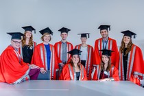 A colection of graduates sitting at/on a table, facing the camera and smiling