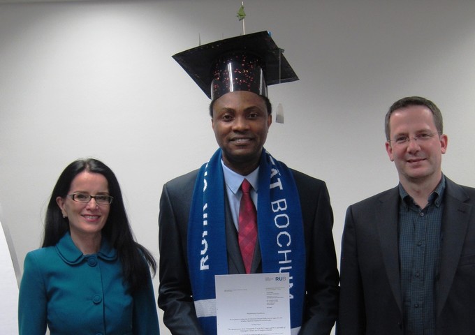 Oluwaseun Fatoba, who is wearing a Ruhr Uni scarf, mortarboard and holding his PhD certificate. On his left is Denise Manahan-Vaughan. On his right is Oliver T. Wolf. All are smiling