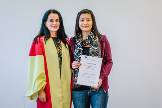 Two women smiling at the camera. One is holding a certificate, the other is dressed in robes