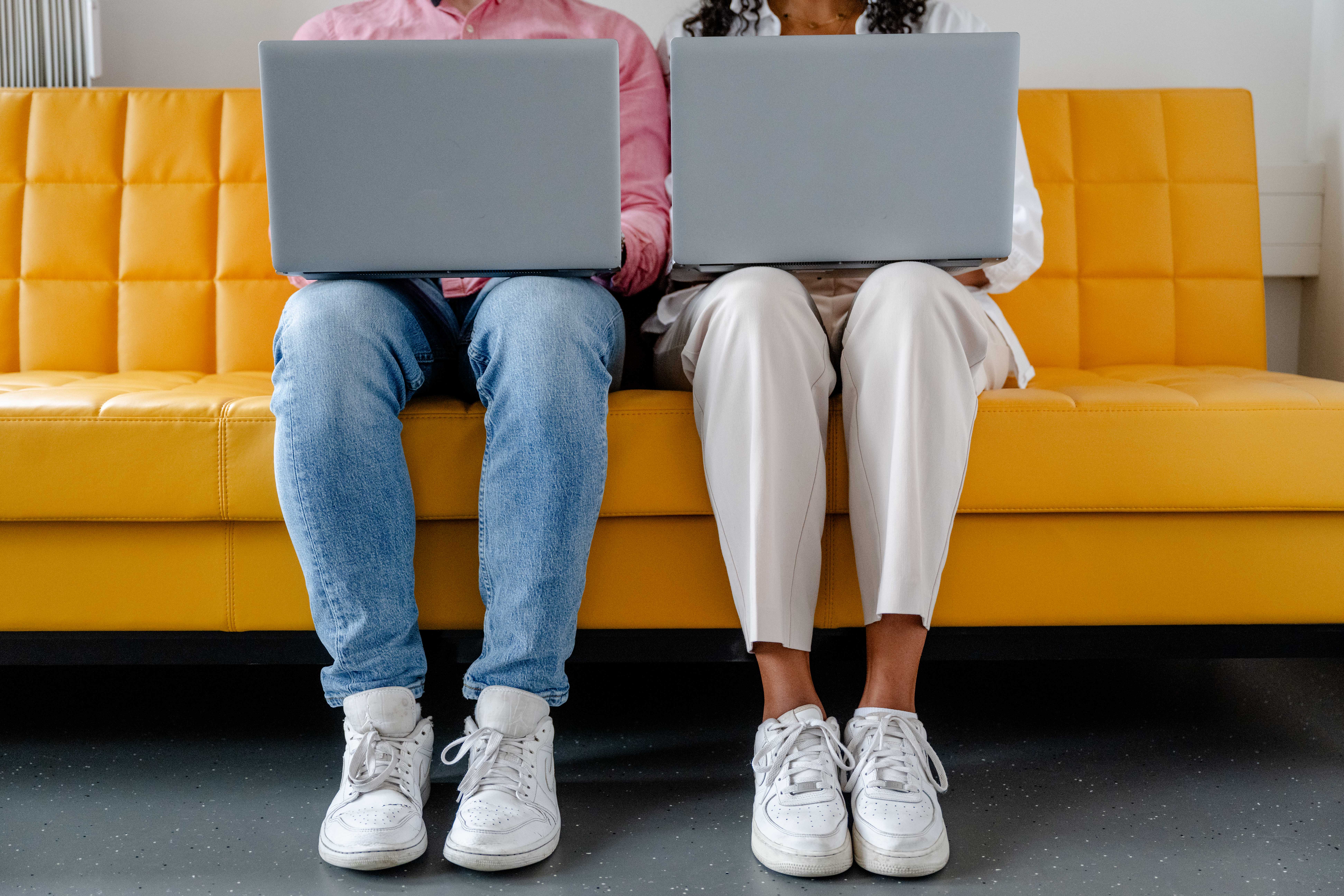 Zwei Personen mit Laptop auf den Knien auf einer gelben Couch