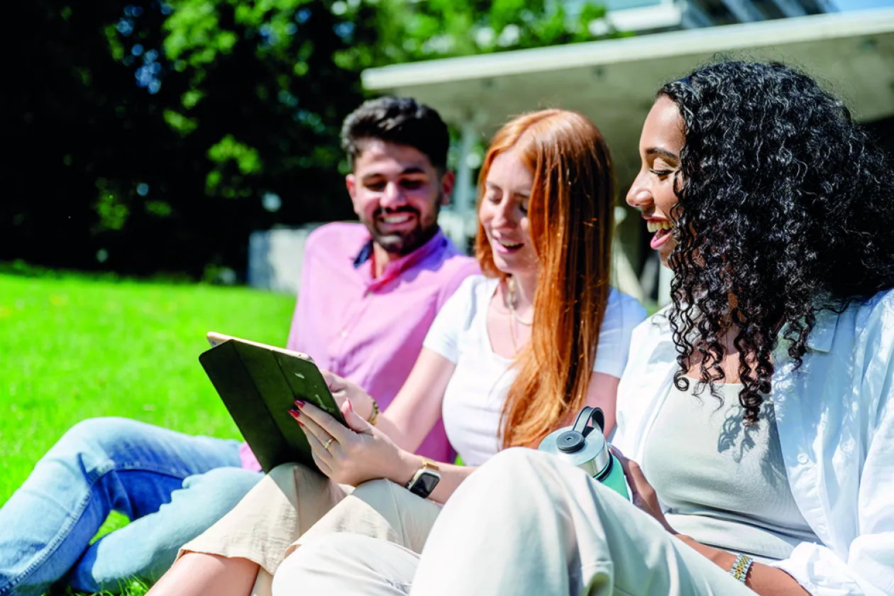 Zwei junge Frauen und ein junger Mann sitzen nebeneinander auf einer Wiese und schauen gemeinsam auf ein Tablet, das die junge Frau in der Mitte in der Hand hält.