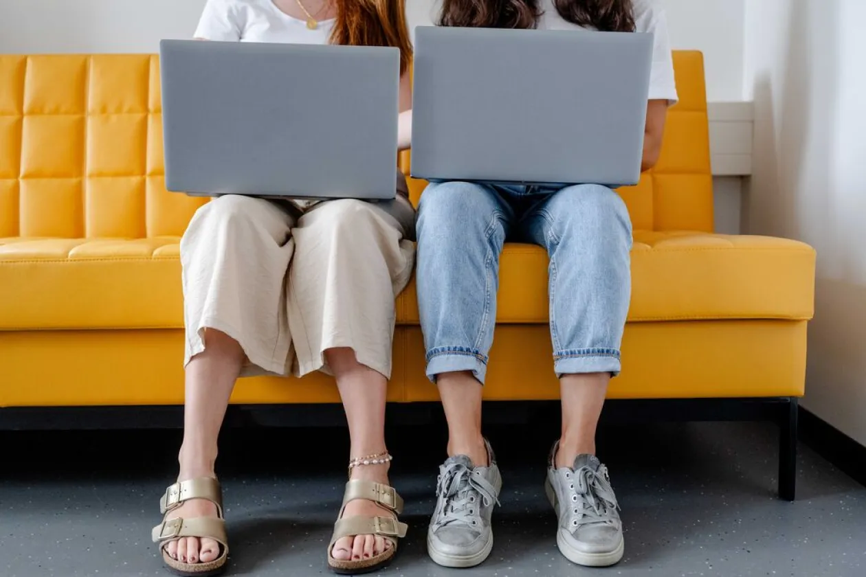 Zwei Frauen sitzen nebeneinander auf einer gelben Couch. Sie haben Laptops auf dem Schoß. 