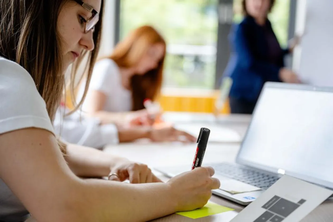Studentin schreib etwas auf. Im Hintergrund ist eine Lehrperson zu sehen. 