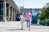 Foto: Zwei Menschen gehen nebeneinander. Sie lächeln. Hinten ist ein Schild. Auf dem Schild steht: Willkommen