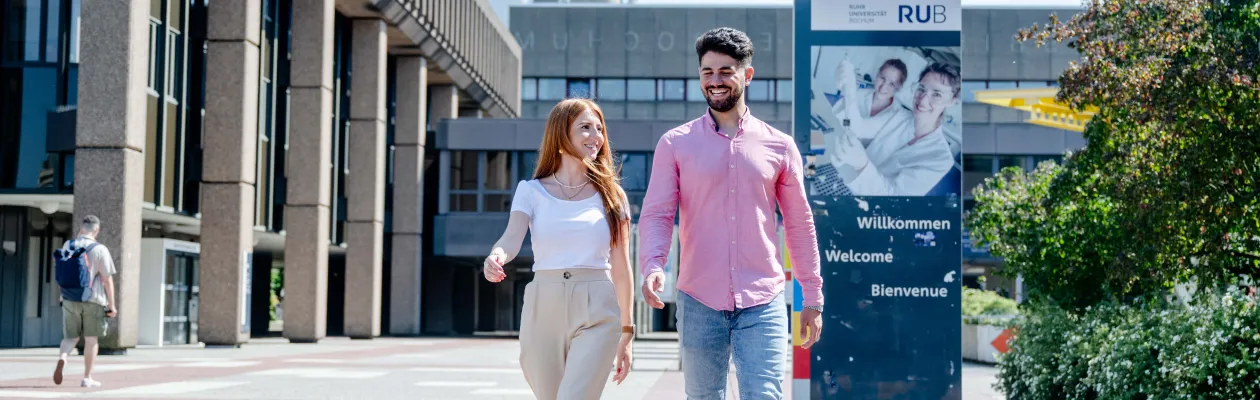 Foto: Zwei Menschen gehen nebeneinander. Sie lächeln. Hinten ist ein Schild. Auf dem Schild steht: Willkommen